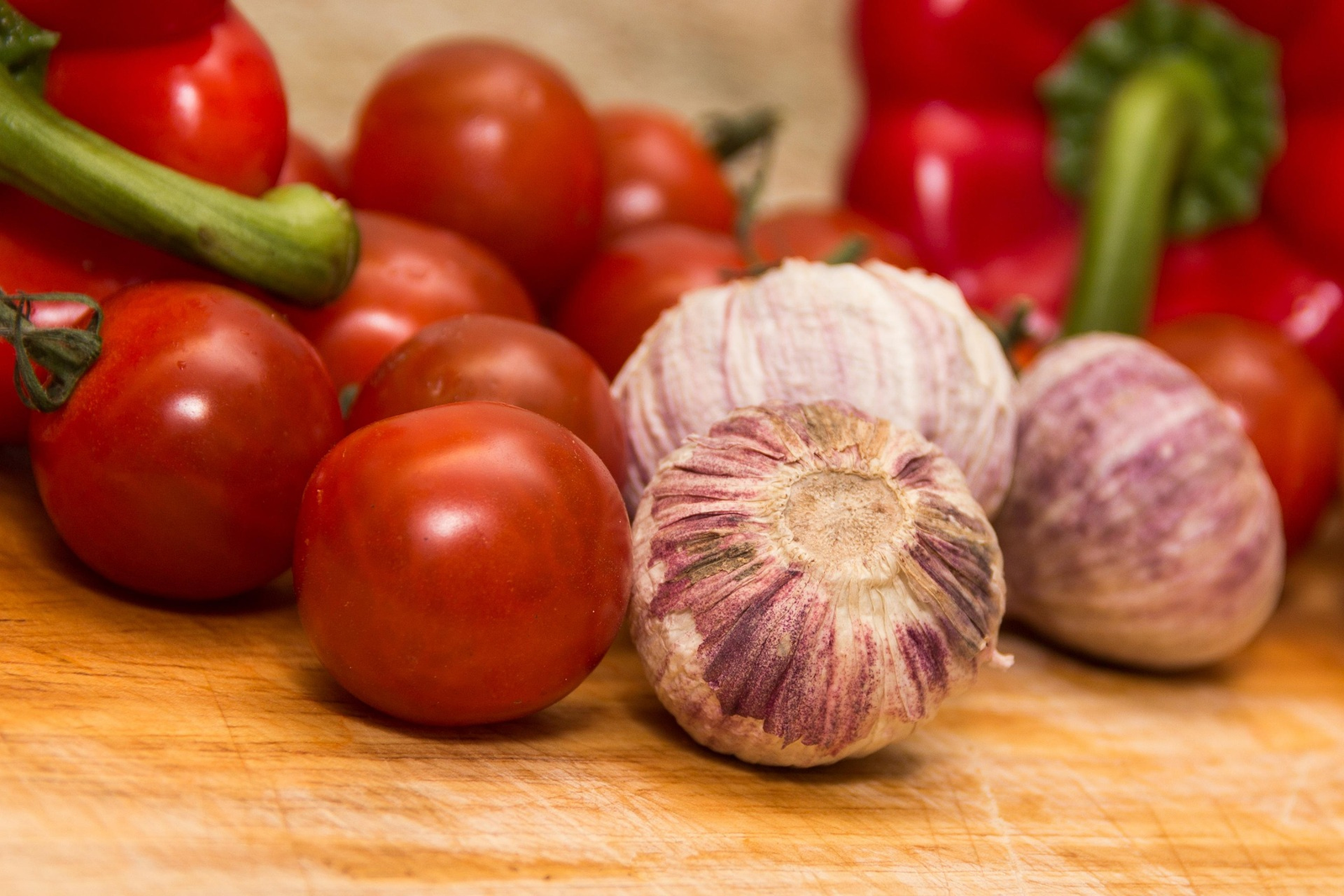 Fresh roma tomatoes, whole garlic bulbs, and red bell peppers piled on a wooden cutting board