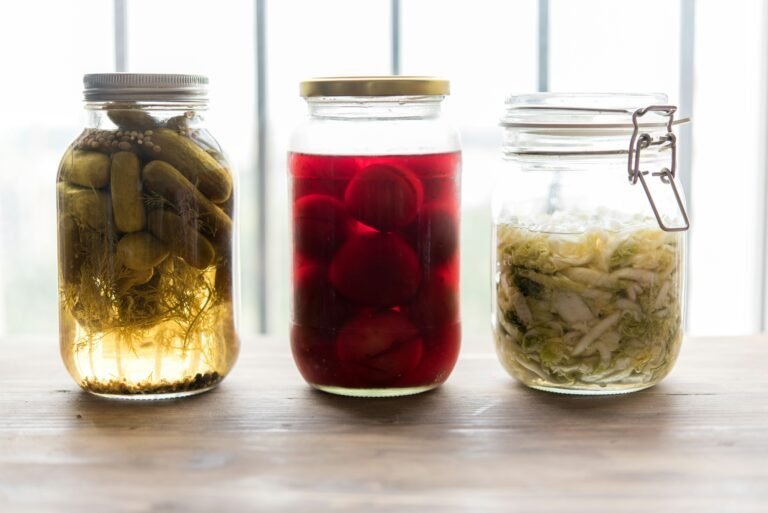 Three glass jars of fermented vegetables — pickles, beets, and sauerkraut — lined up on a wooden counter