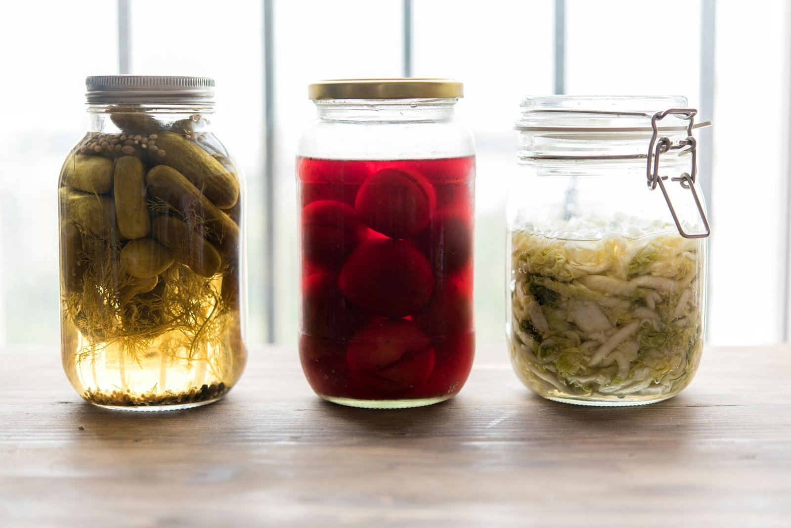 Three glass jars of fermented vegetables — pickles, beets, and sauerkraut — lined up on a wooden counter